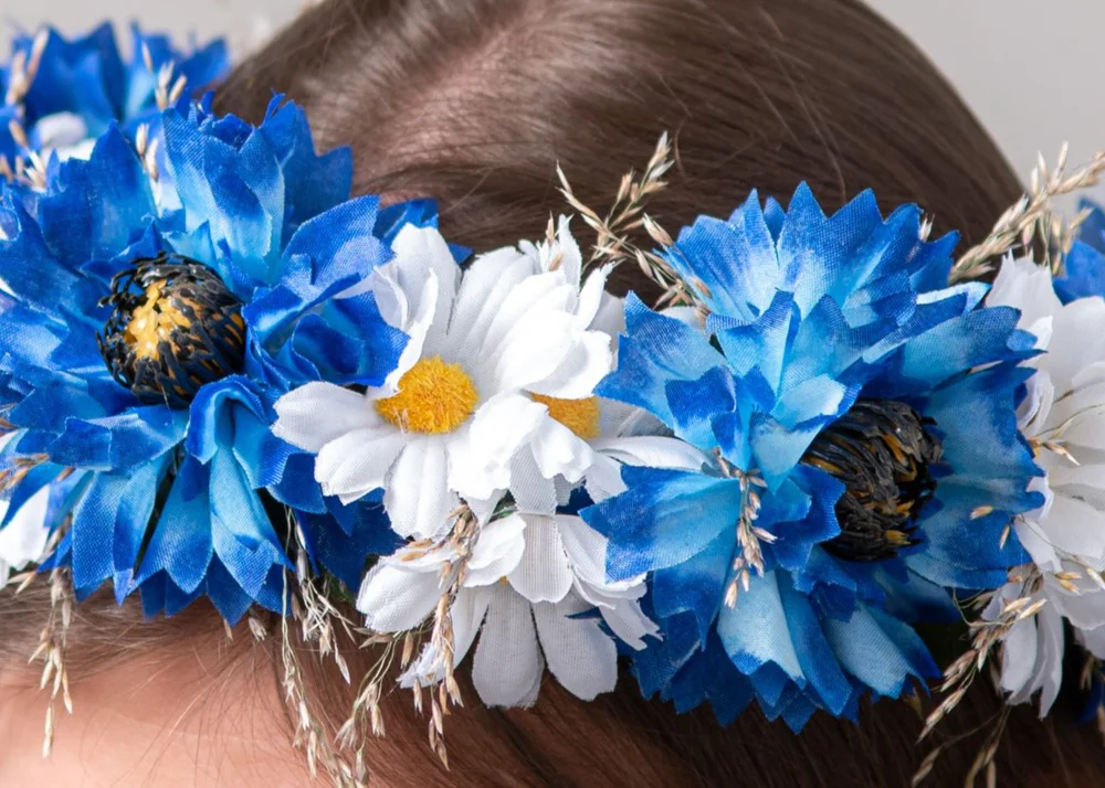 Bilde tuvplānā: mākslīgs vainags ar zilām rudzupuķēm un baltiem ziediem, un dabīgām smilgām